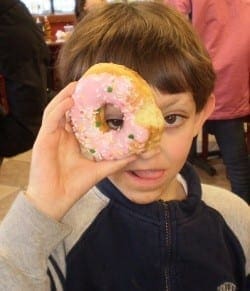 A young boy holds a pink frosted donut with sprinkles in front of his eye.