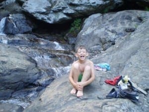 Young boy sitting on rocks by a waterfall with clothes and backpack nearby.