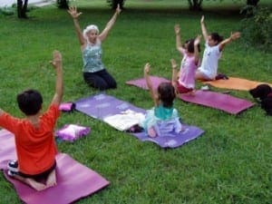 Adult and children practice yoga outdoors on mats with arms raised.