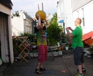 Two boys in an outdoor recreational area, one lifting a medicine ball overhead while the other watches.