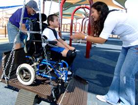 A boy in a wheelchair is on a playground swing with two adults helping him.