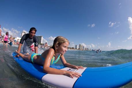 A young girl lies on a surfboard as a man paddles behind her in the ocean with buildings in the distance.