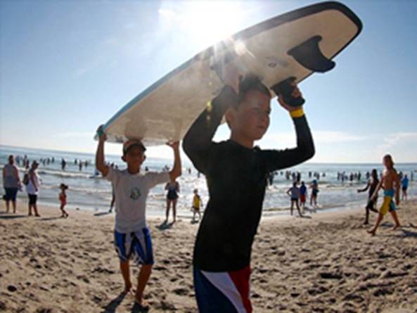 Two children carry a surfboard on a beach with the ocean and spectators in the background.