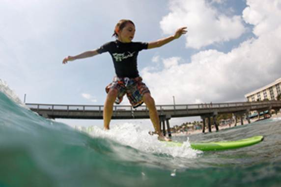 A child surfs a wave with a pier in the background.