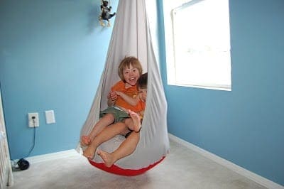 Child with a bright orange shirt sits in a hanging swing chair in a room with light blue walls.
