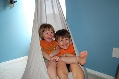 Two smiling children in orange shirts sit in a white fabric swing.