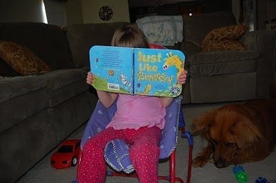 Child sits in a chair reading a book with a dog nearby.