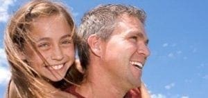 Father smiling while giving his daughter a piggyback ride against a clear blue sky.