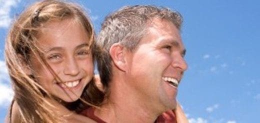 Father smiling while giving his daughter a piggyback ride against a clear blue sky.