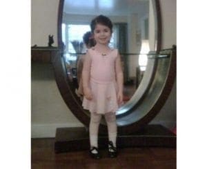 Child in ballet attire smiles in front of a large mirror.
