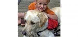 A smiling child rests his head on the back of a Labrador Retriever wearing a red vest.