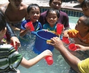 Children play with cups and a bucket in shallow water.