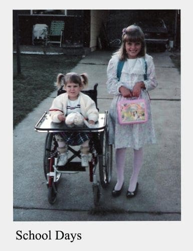 Two children stand on a sidewalk, one in a wheelchair and one holding a lunchbox.