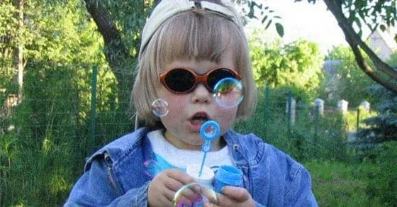 Child wearing sunglasses and a hat blowing bubbles outdoors.