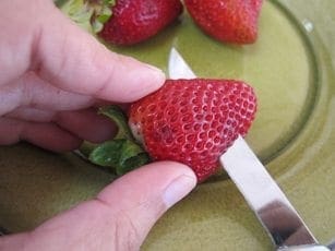 A hand holds a strawberry while a knife cuts off the top.