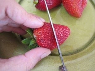 A hand holds a strawberry while a knife cuts into it.