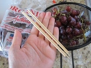 Hand holding wooden skewers next to grapes and a bag of raisins.