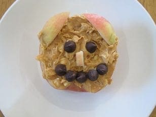 Apple half with peanut butter, chocolate chip face, and apple slice ears on a white plate