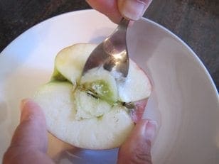 Hands core an apple half with a fork over a white bowl to make a scary face.