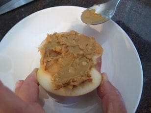 Hands holding an apple half while a spoon spreads peanut butter on it.