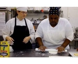 Two bakers in a kitchen, one rolling out dough.