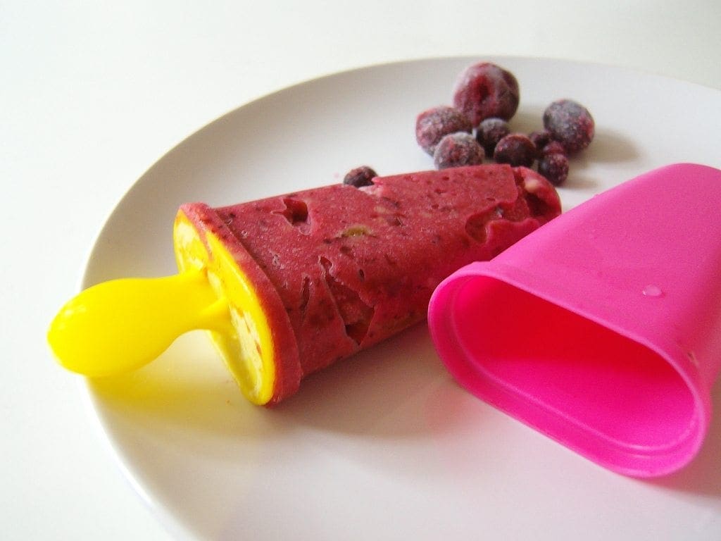 Fruit popsicle with a yellow handle and a pink cap next to frozen berries on a white plate.