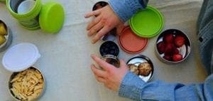 Hands packing various food items into round metal lunch containers with colorful lids.