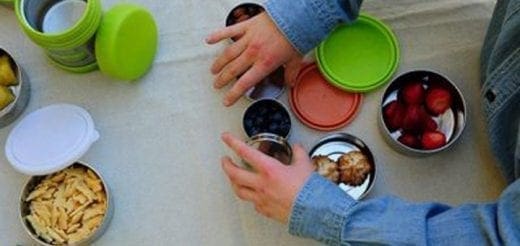 Hands packing various food items into round metal lunch containers with colorful lids.