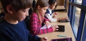 Three children sit at a desk using tablets by the window.