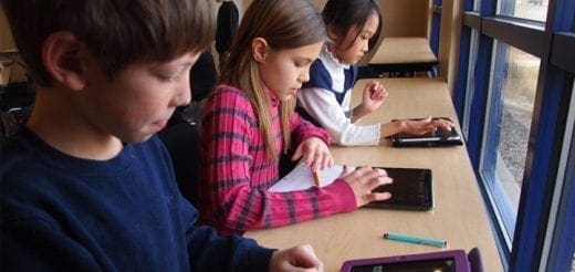 Three children sit at a desk using tablets by the window.