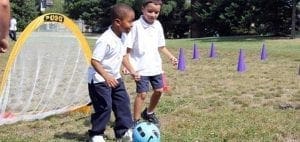 Two boys in white polo shirts play soccer with a pop-up goal and purple cones.