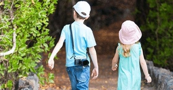 Two children walk down a path with lush greenery.