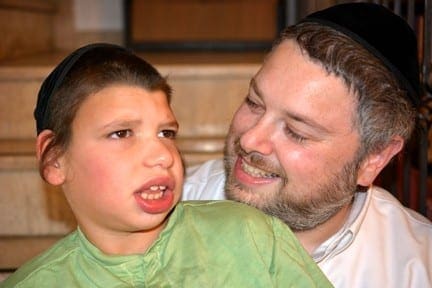 A man with a beard smiles at a boy wearing a black yarmulke and a green shirt.