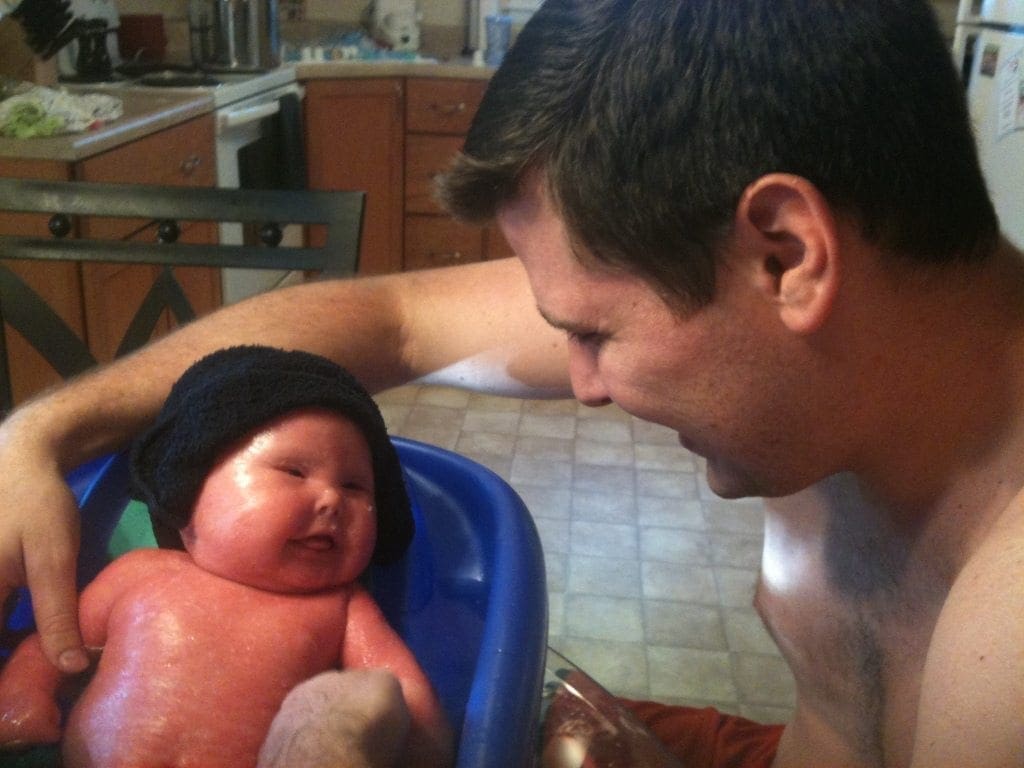 A man looks down at a baby in a blue tub, both wearing black hats.
