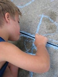 Young boy focused on drawing shapes with blue chalk on a concrete driveway as a summer activity.