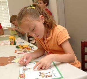 Young girl focused on a coloring worksheet while sitting at a table with other children in a classroom.