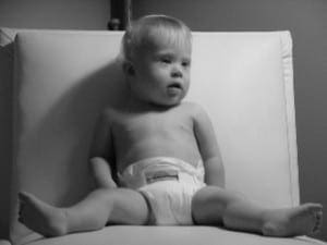 Jacob, a baby with Down Syndrome, sits upright on a white chair while looking away from the camera.