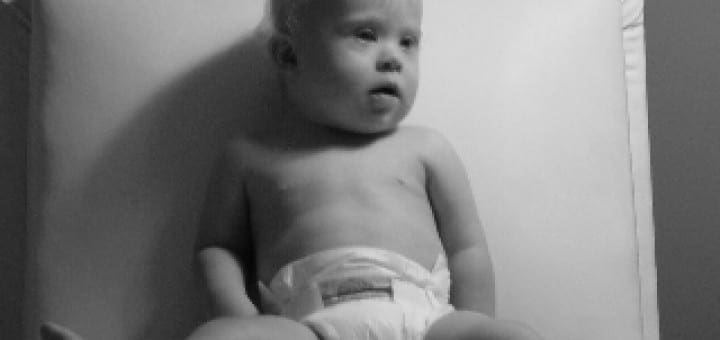 Jacob, a baby with Down Syndrome, sits upright on a white chair while looking away from the camera.