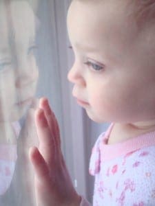Young toddler pressing her hand against a window while curiously gazing at her own reflection.