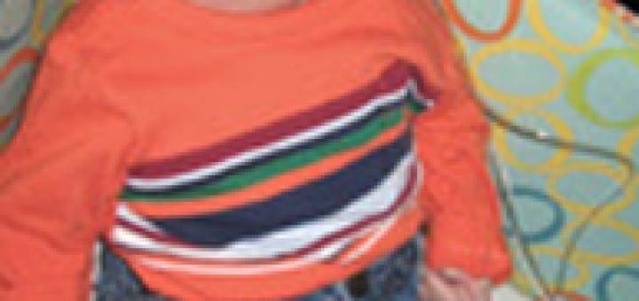 Eliot Mooney sits in his crib wearing an orange striped shirt and blue jeans while using a nasal cannula.