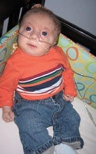 Eliot Mooney sits in his crib wearing an orange striped shirt and blue jeans while using a nasal cannula.