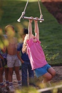 Happy child in a pink shirt swinging from playground monkey bars while friends watch nearby.