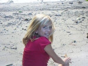 Happy young girl with blonde hair wearing a red shirt, smiling while sitting on a beach.