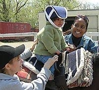 Young child wearing a protective helmet while riding a horse supported by two adults during a therapy session.
