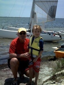 Wyatt wearing a life jacket stands next to his father Jim in front of a sailboat at Sebastian Inlet State Park.