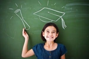 Smiling girl stands in front of a chalkboard with chalk drawings of a graduation cap and diploma.