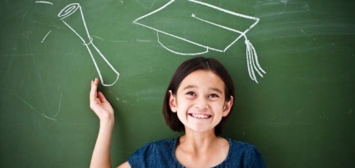 Smiling girl stands in front of a chalkboard with chalk drawings of a graduation cap and diploma.