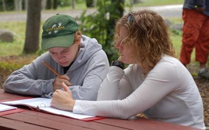 Two people sit at a picnic table with notebooks and pencils.