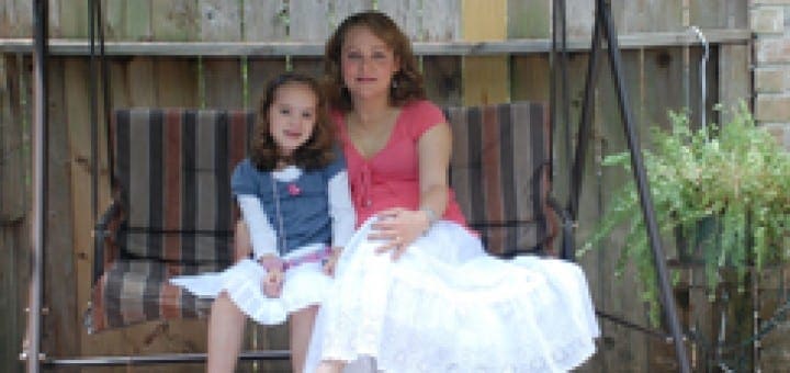 Two females sit on a swing set in front of a wooden fence.