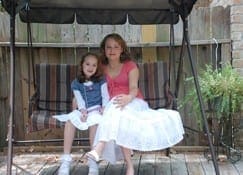 Two females sit on a swing set in front of a wooden fence.
